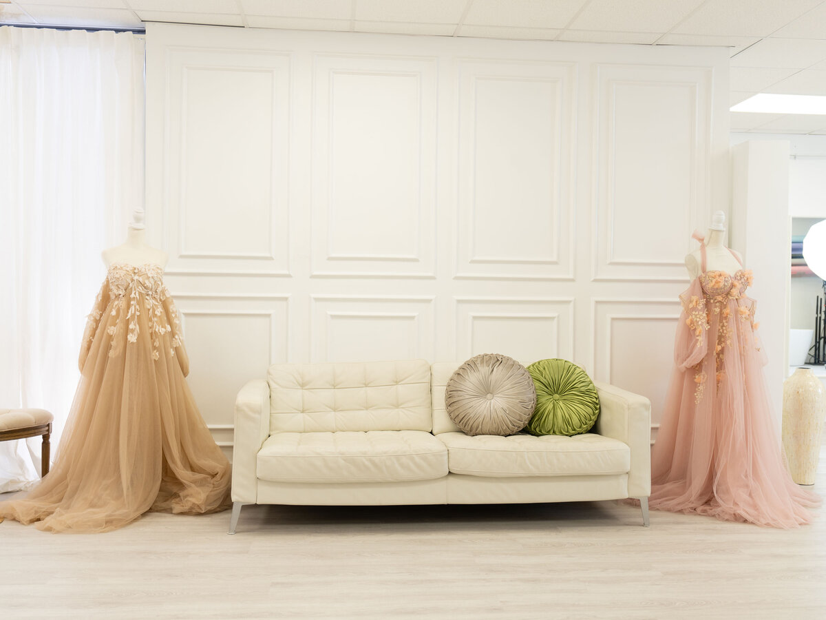 A beige sofa with two round pillows, one gray and one green, sits against a white paneled wall. Two mannequins in elegant, flowing gowns flank the sofa.