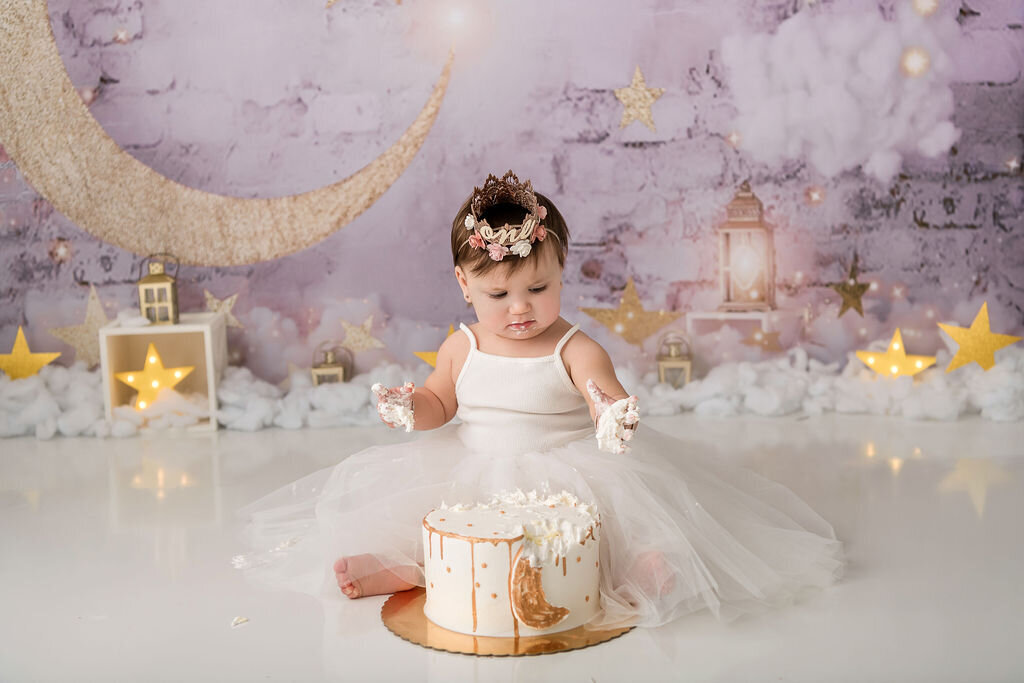 Baby girl wearing floral crown explores white and gold drip cake during whimsical moon and stars first birthday photoshoot