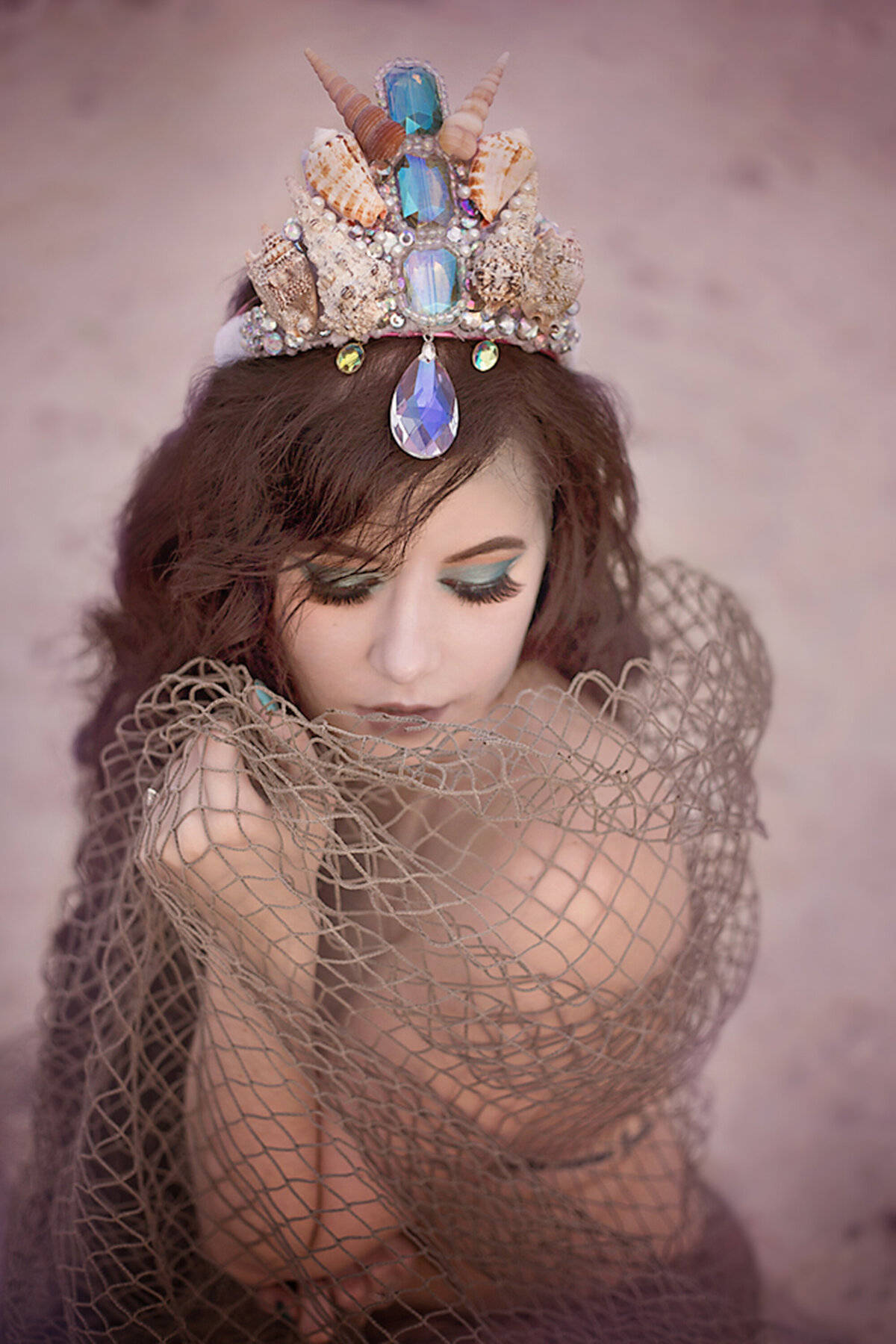 Adult woman with long brown hair and mermaid crown with shells and jewels.  Her eyes are closed and she is wearing green eyeshadow and thick eyelashes.  The is wrapped in a fishing net.