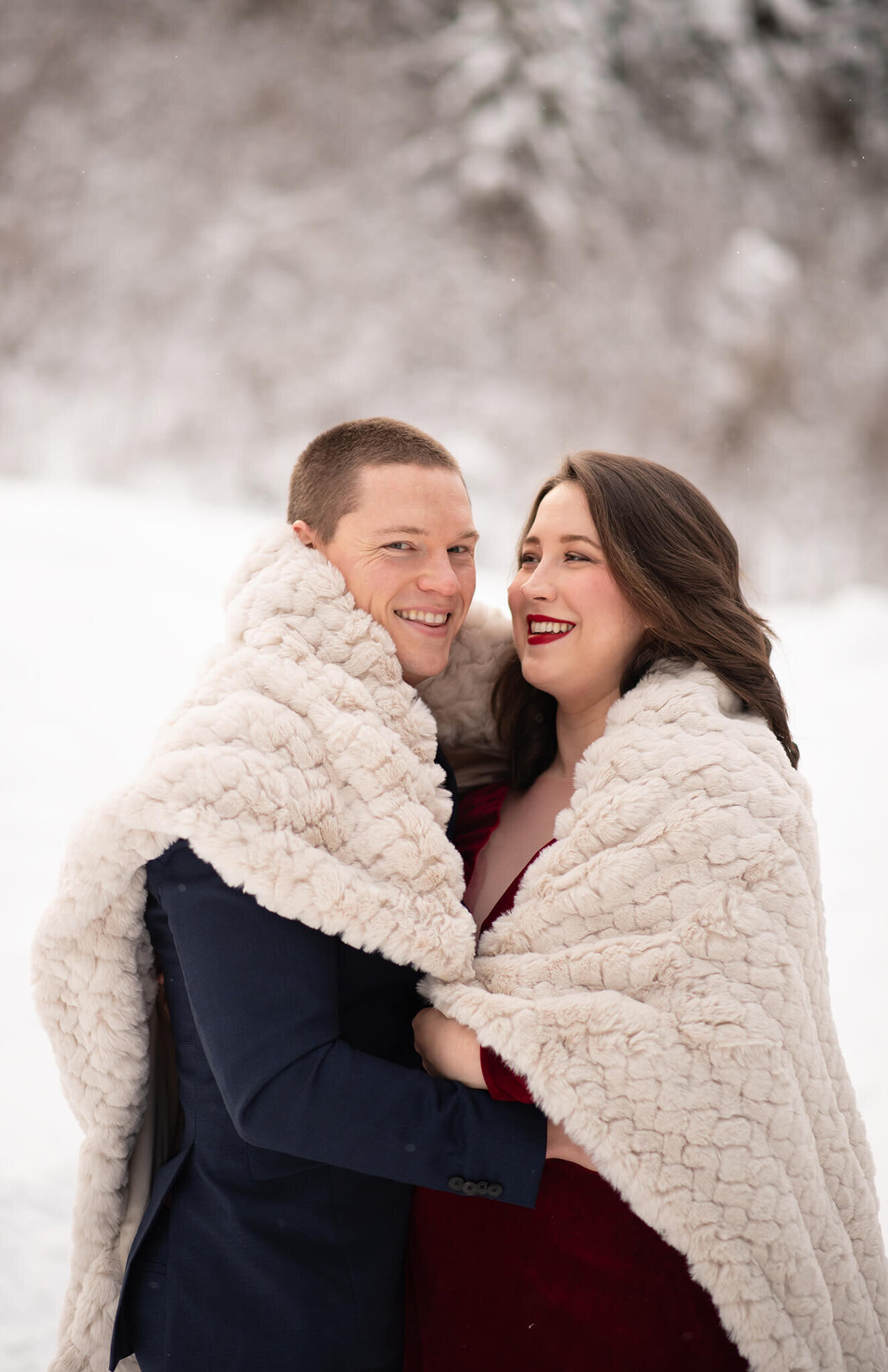 A husband and wife are in a cozy embrace under a soft blanket. Their faces are close together and the wife smiles at her husband while he looks at the camera. There is a snowy backdrop. Taken by Brittany Danielle Photography