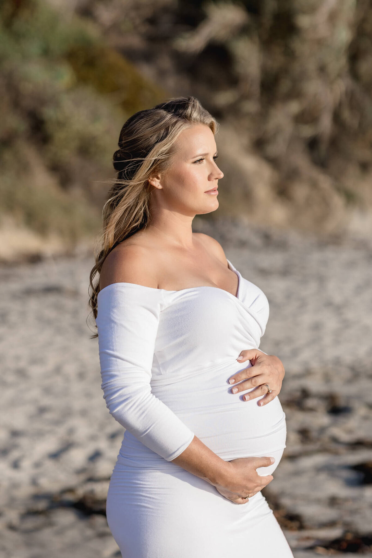 A pregnant woman in a white off-shoulder dress stands on a beach, gently holding her belly. She gazes into the distance with a serene expression. The background features sand and blurred greenery.