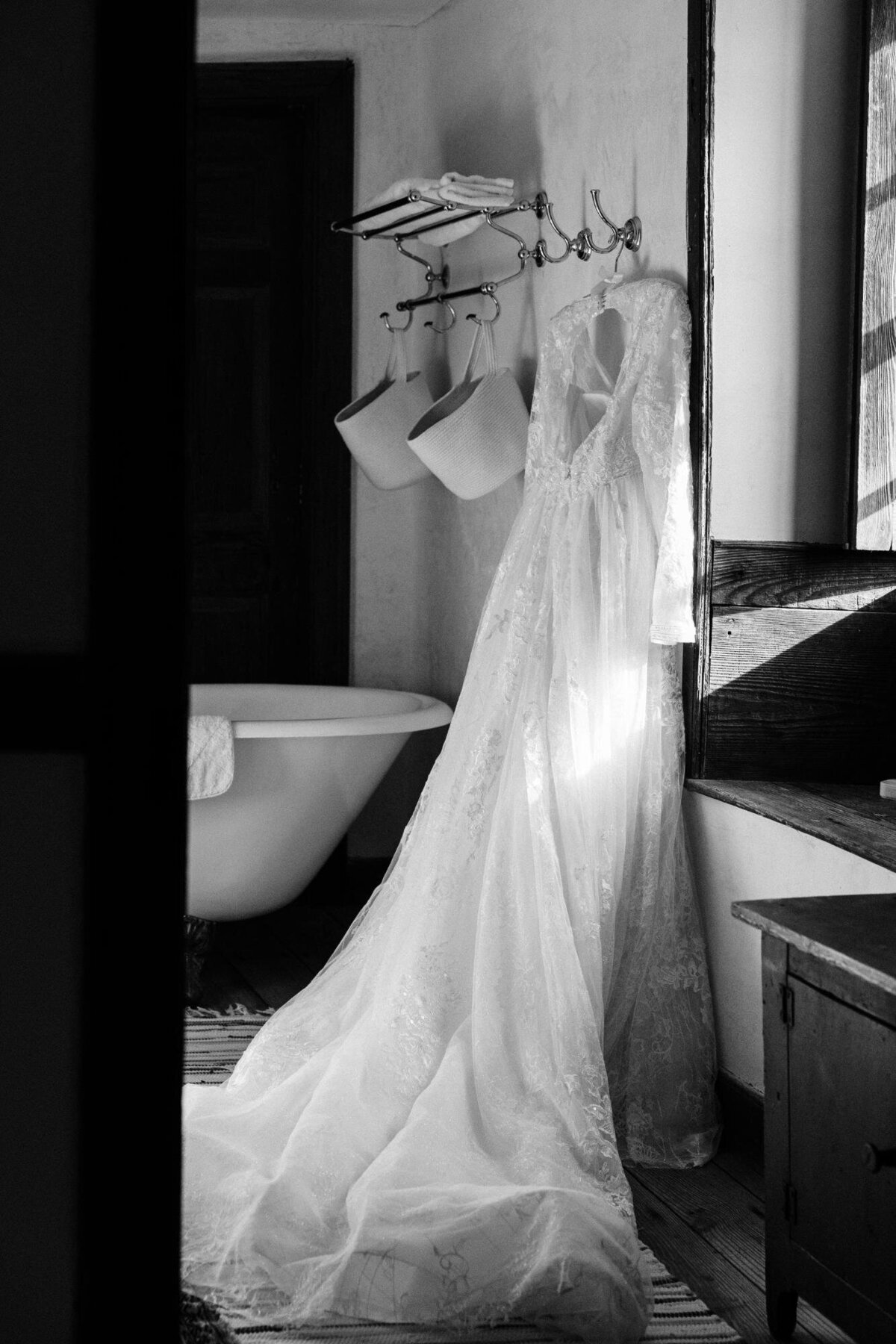 A lace wedding dress hanging beside a bathtub in a room with natural light casting shadows.