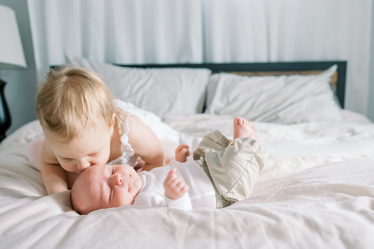 A blonde young toddler gives her new infant brother a kiss on the cheek