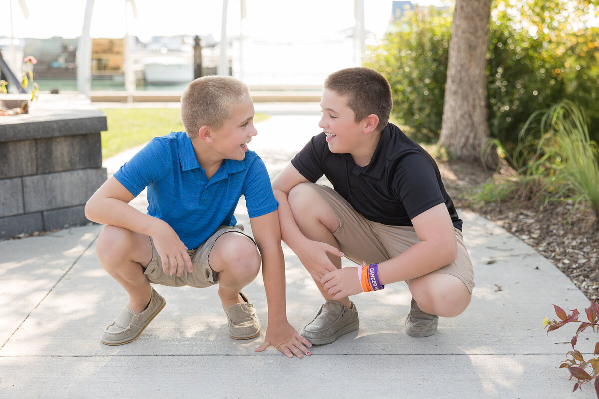 Twin brothers are crouching down on a sidewalk and smiling at each other