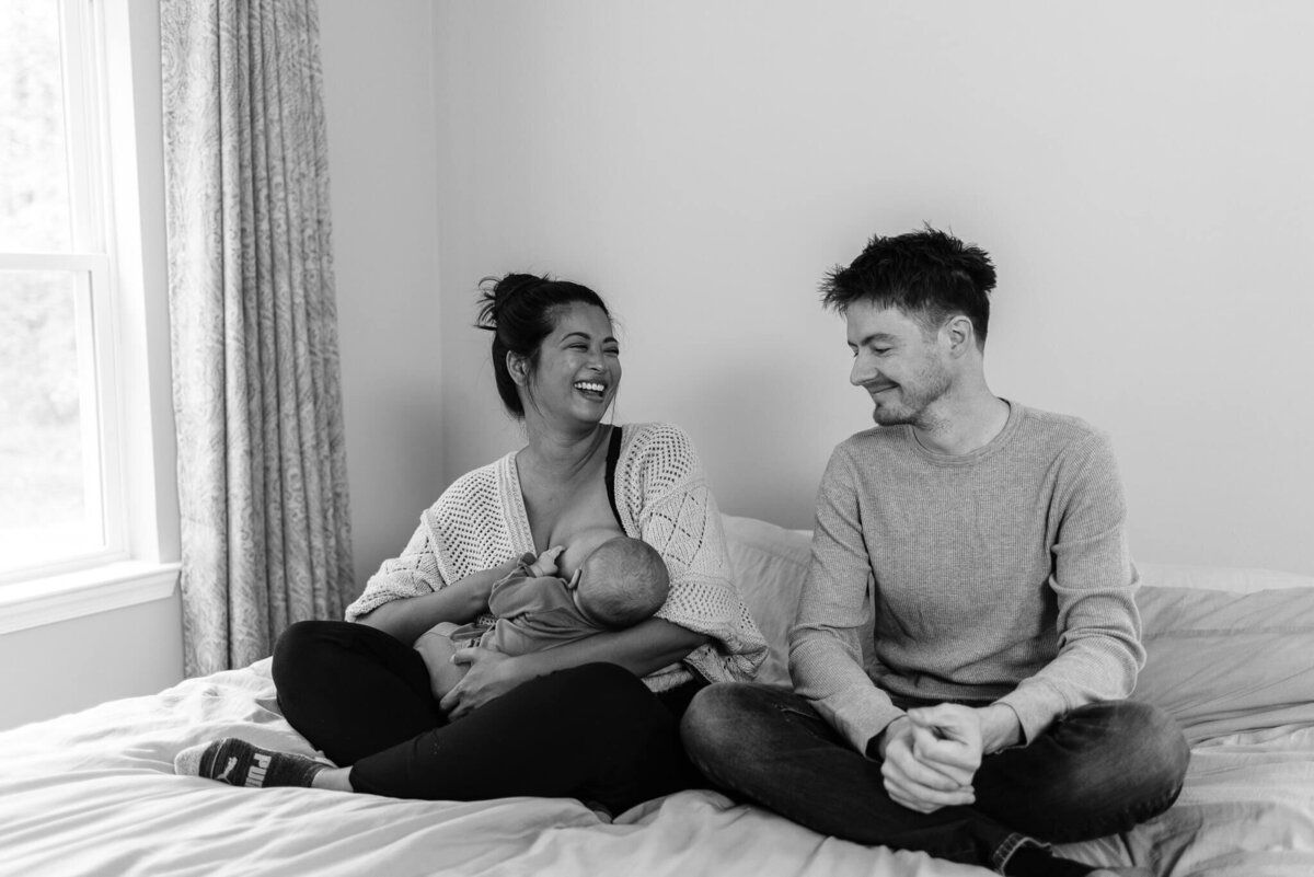 B&W photo of a breastfeeding mom laughing at her husband in bed
