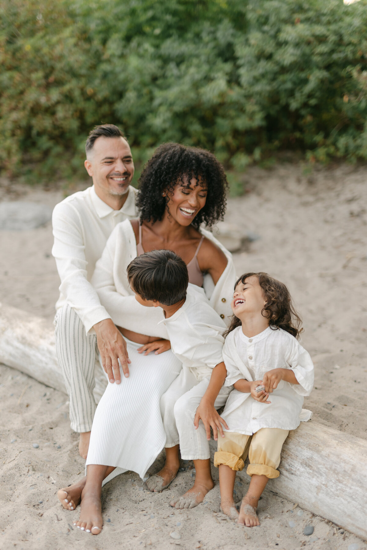 Mom and Dad are laughing at their smiling boys while sitting at a log in a Toronto beach