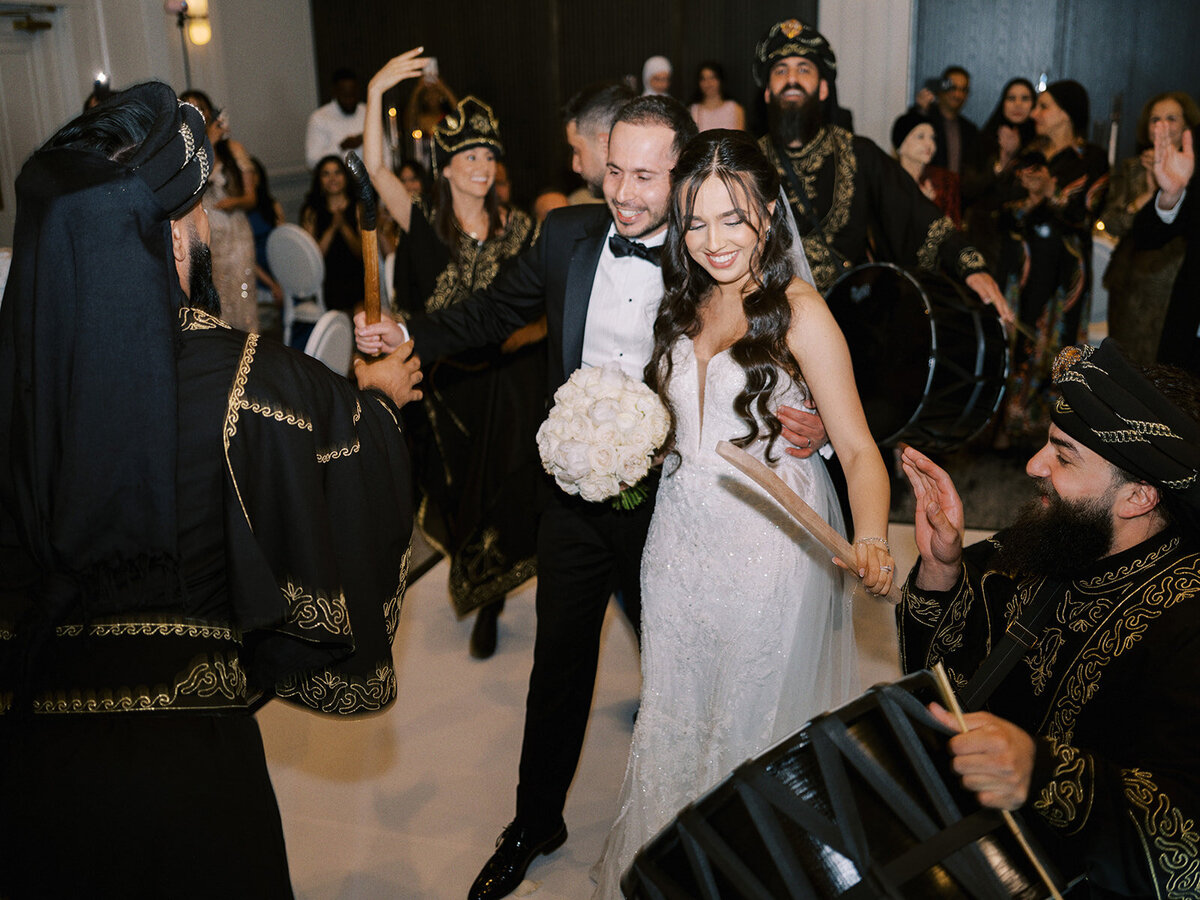 A bride and groom in formal attire celebrate their classic Calgary wedding by dancing with a group of performers wearing traditional costumes, with musicians playing drums.