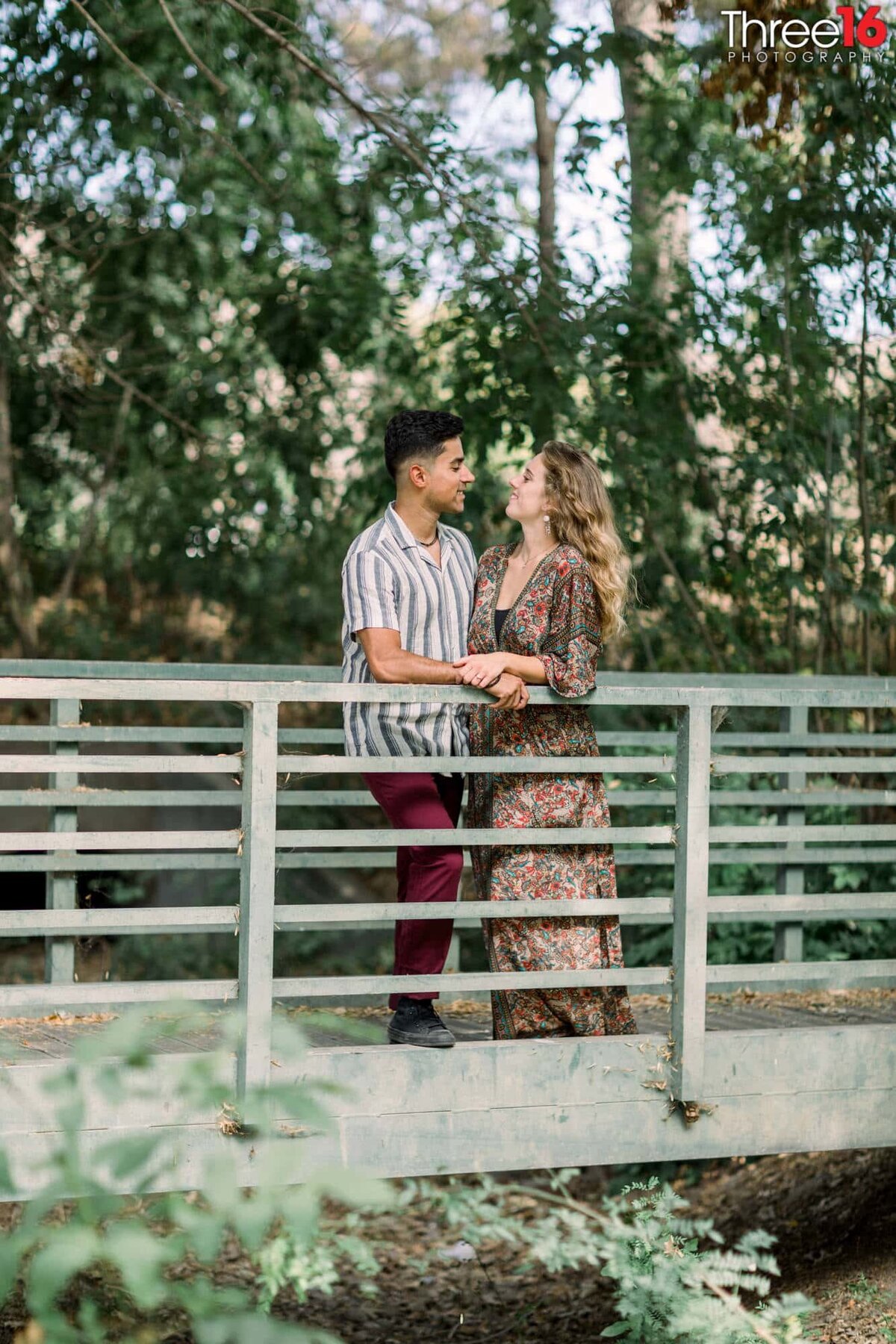 Engaged couple stand together against a bridge railing gazing into each other's eyes