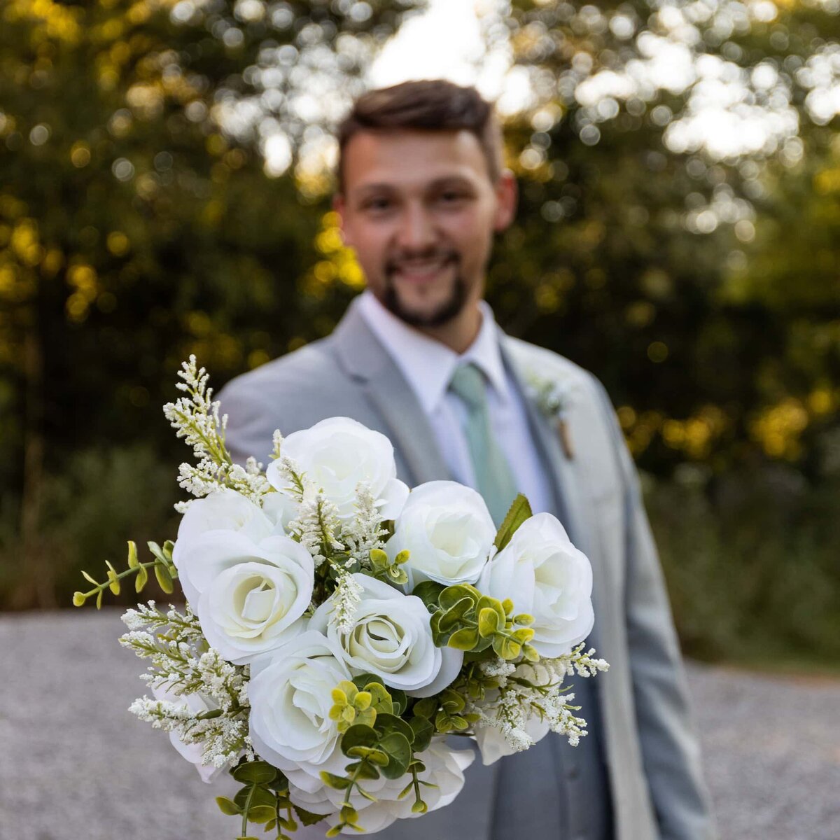 Groom holding bride’s bouquet out at camera in a role reversal photo featuring the focus on the flowers and shallow depth of field resulting in the groom being soft and the beautiful boka in the background