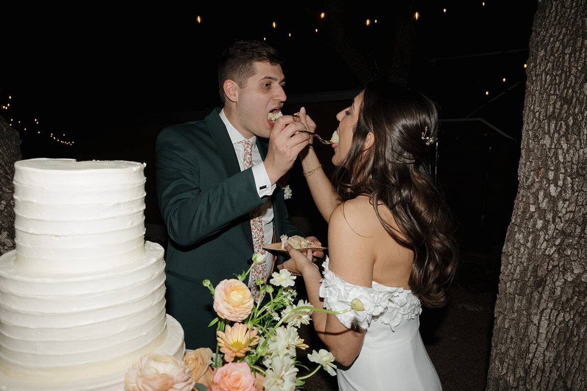Bride and groom feeding eachother cake. In front of their cake and flowers. 
