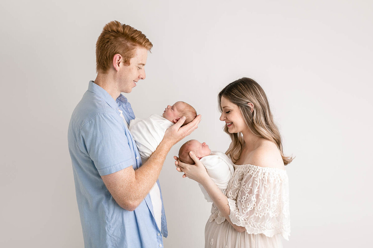 Image of Mom and Dad holding twins facing one another. Family is wearing light neutral tones and colors. Image taken at Portland Oregon newborn photography session at SE Portland newborn studio.