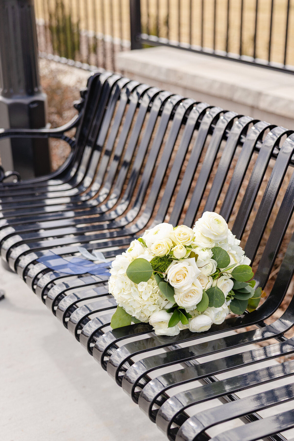 A beautiful white wedding bouquet with roses and eucalyptus rests on a black metal bench