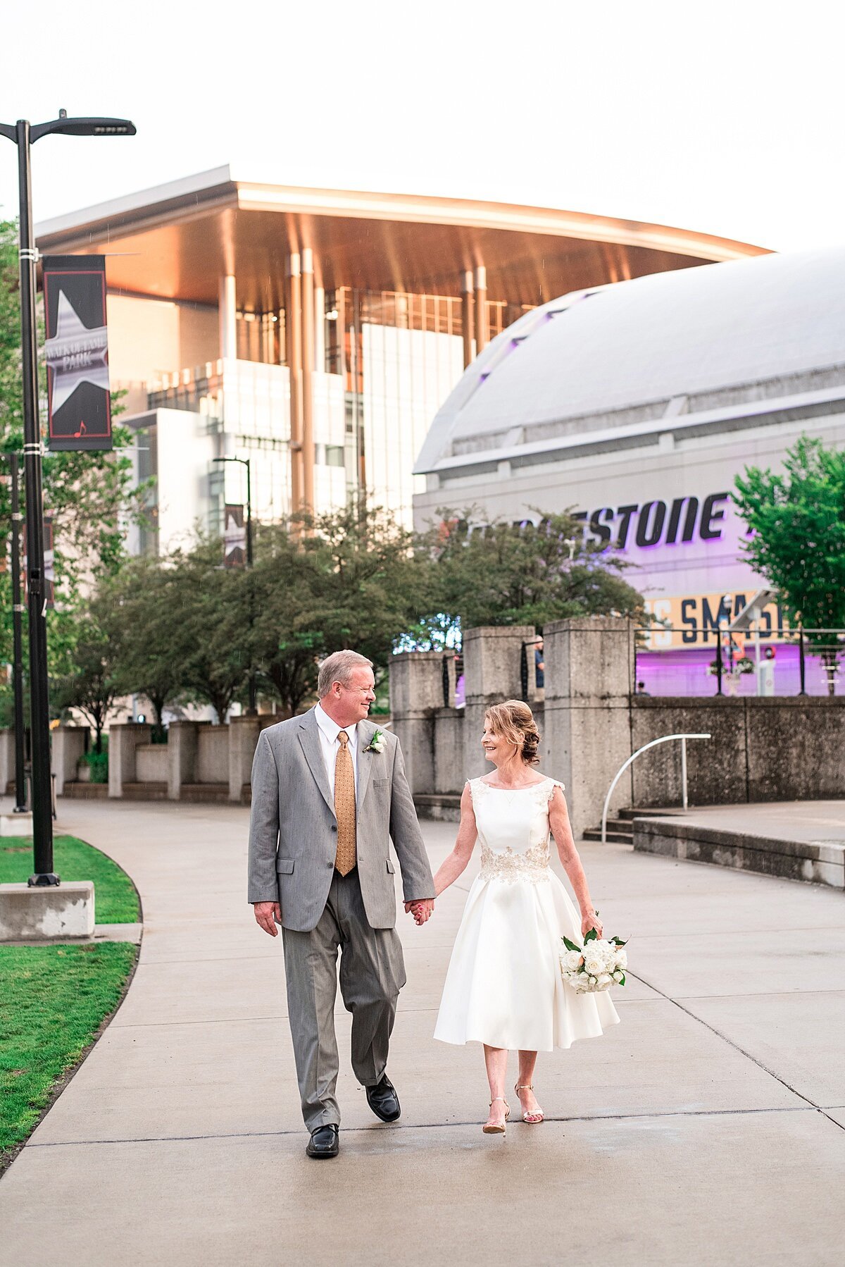 The bride and groom walk passed the Smashville sign above Bridgestone Arena were the Preds play in Nashville. They are holding hand and looking at each other. The groom is wearing a gray suit with a white shirt and pink tie. The bride is wearing a white tea length wedding dress with gold lace detail around the waist and at the shoulders. She is carrying a small white bouquet at her side.