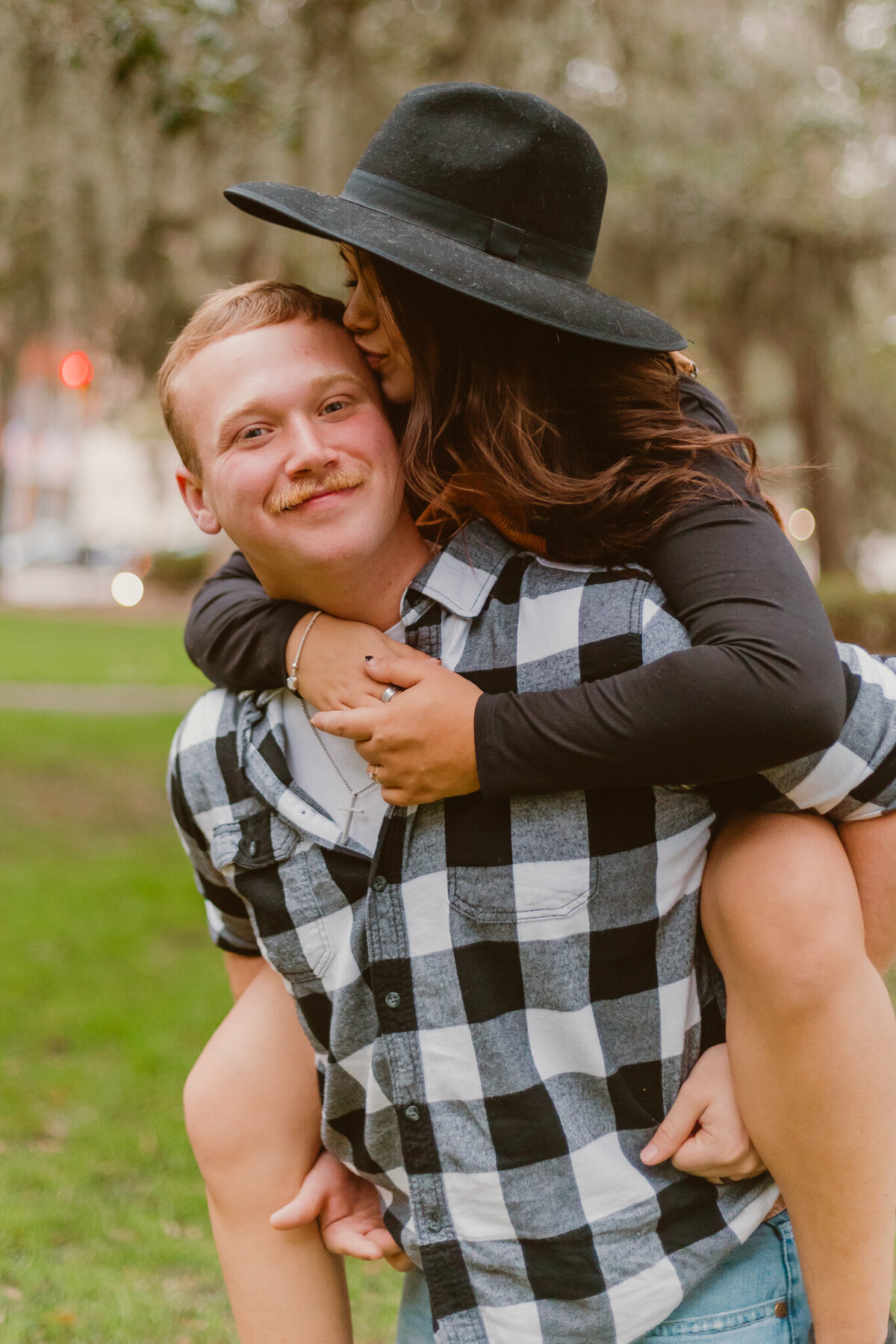 Couple sharing a playful piggyback ride in a park in Savannah, Georgia. The woman, wearing a dark hat, kisses the man on the cheek while he smiles, dressed in a plaid shirt. Captured during a couple’s engagement session by texas photographers ShutterUp Photography.