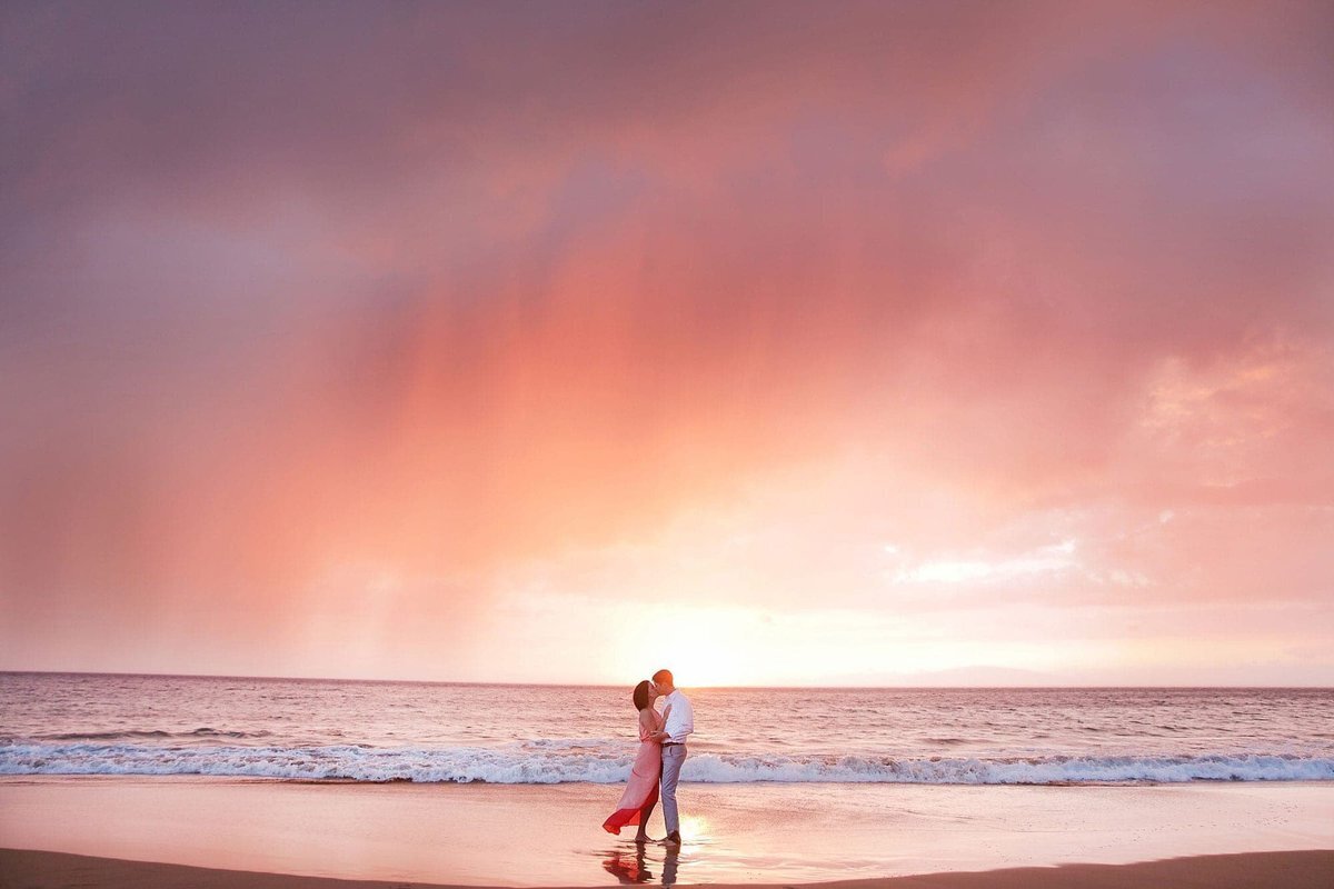 Woman wearing a pink dress being blown by the tradewinds kisses her husband on the beach in south Maui