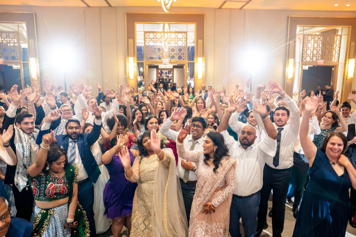 A large group of people dressed in formal attire are dancing and raising their hands joyfully at an indoor event. The background features elegant decor with bright lights and patterned walls.