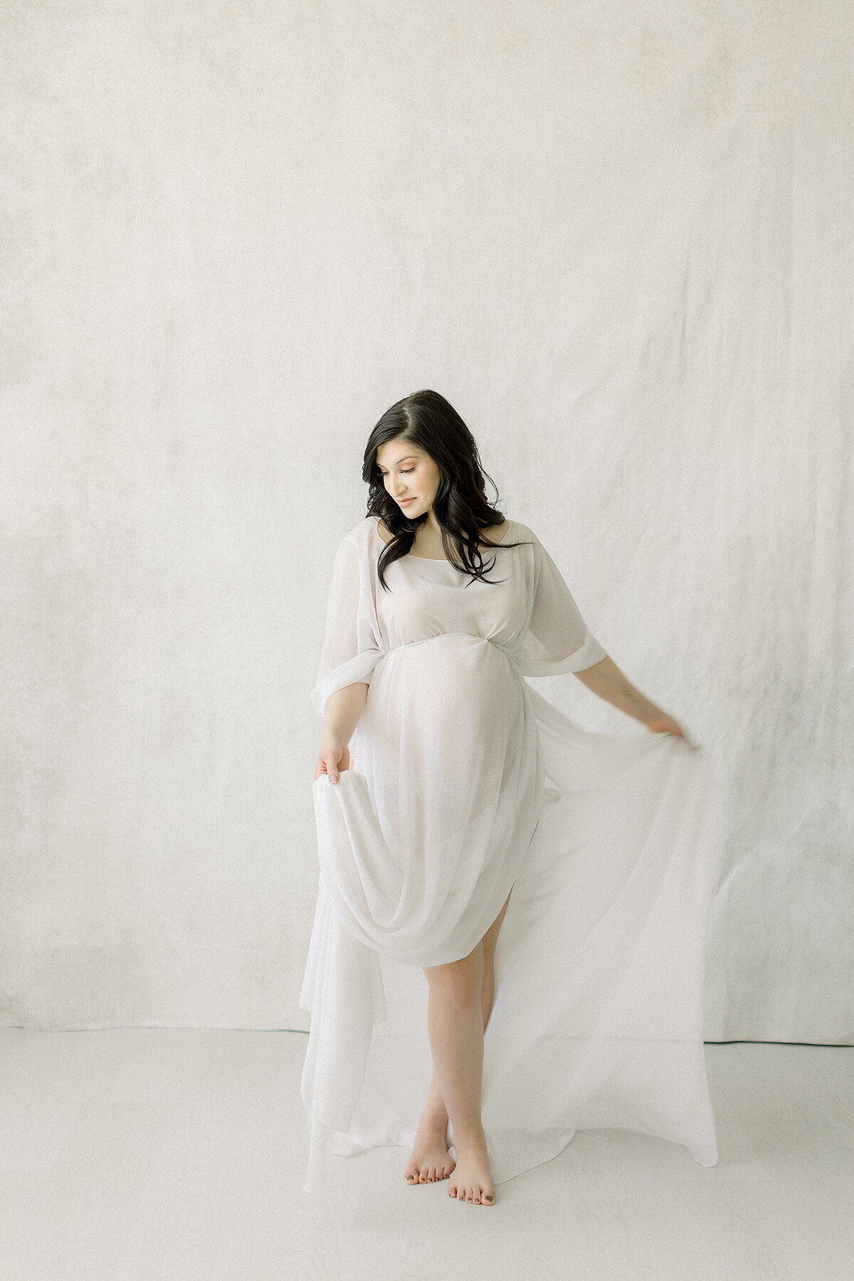 A light filled in studio maternity session taken in a Dallas/Fort Worth area photography studio of an expecting mother while she is playfully holding her chiffon maternity dress.