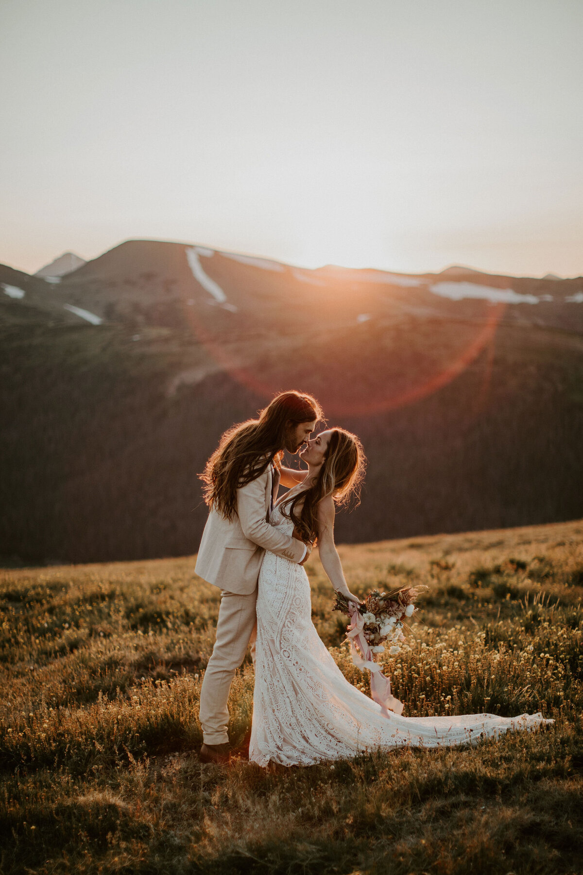 Bride and groom wearing an ivory suit and white wedding gown, kissing during golden hour in a mountain landscape.