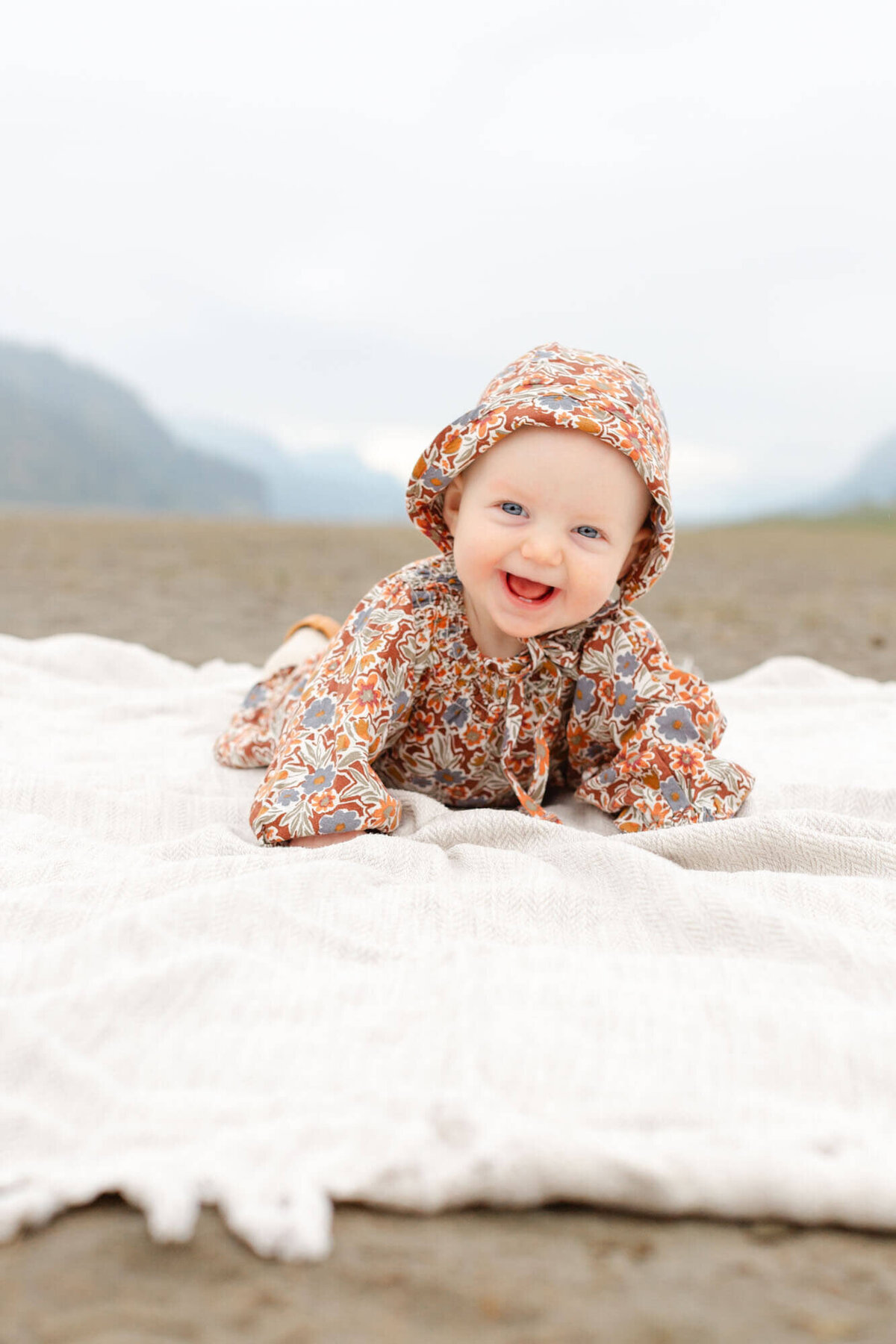 6 month old baby girl in floral romper laying on a cream blanket in the sand with mountain views behind her. She is at Rooster Rock State Park for her milestone photo session in the Columbia River Gorge.