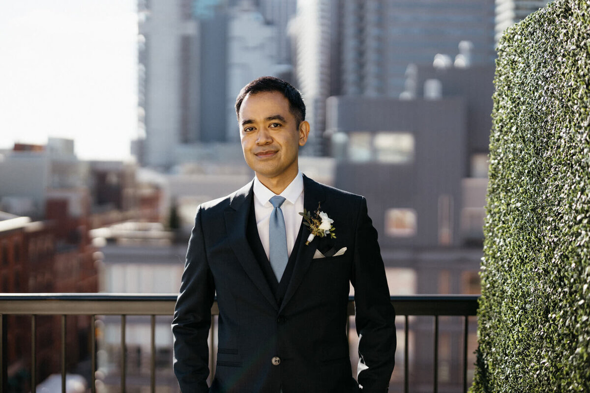 The groom, who is wearing a black suit and tie, is standing on a terrace with buildings in the background.