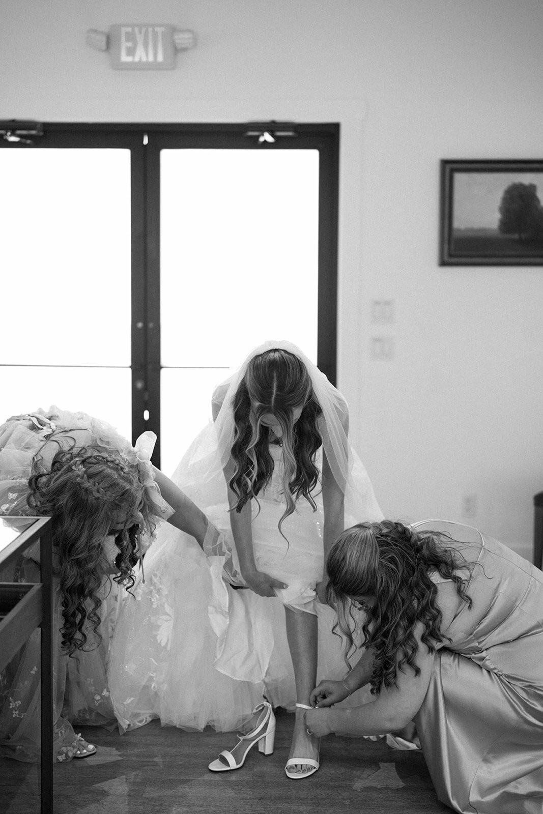 Black and white candid photo of the bride’s mom and sister helping her put on her shoes before the ceremony.