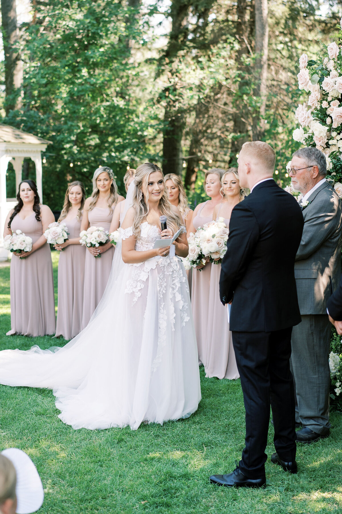 A bride and groom stand facing each other during a modern and romantic wedding in Calgary at Norland Historic Estate. The bride holds a microphone. Bridesmaids in beige dresses stand in a row holding bouquets, while an officiant stands nearby, ready to lead the ceremony.