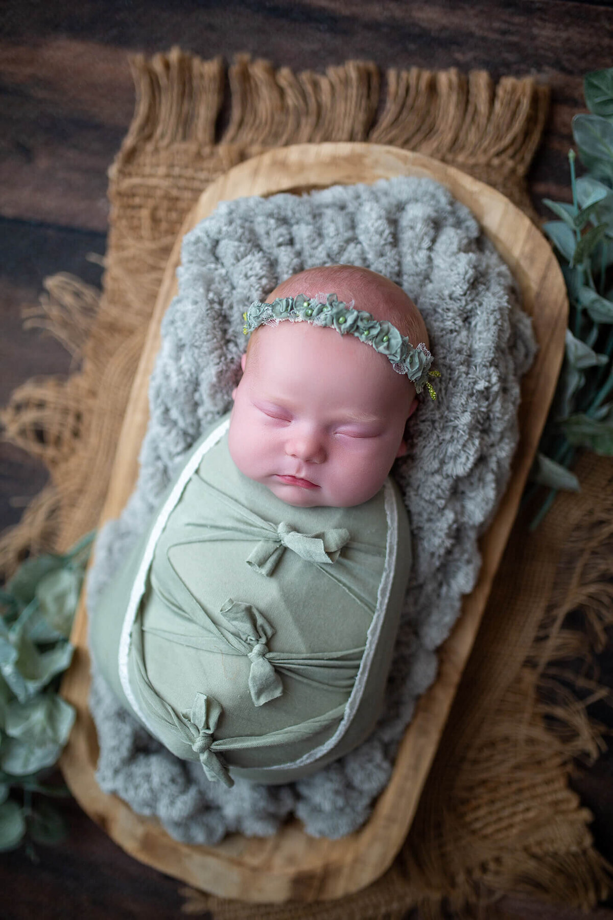 Newborn baby girl wrapped in sage green and bows laying in a rustic bowl, burlap and greenery