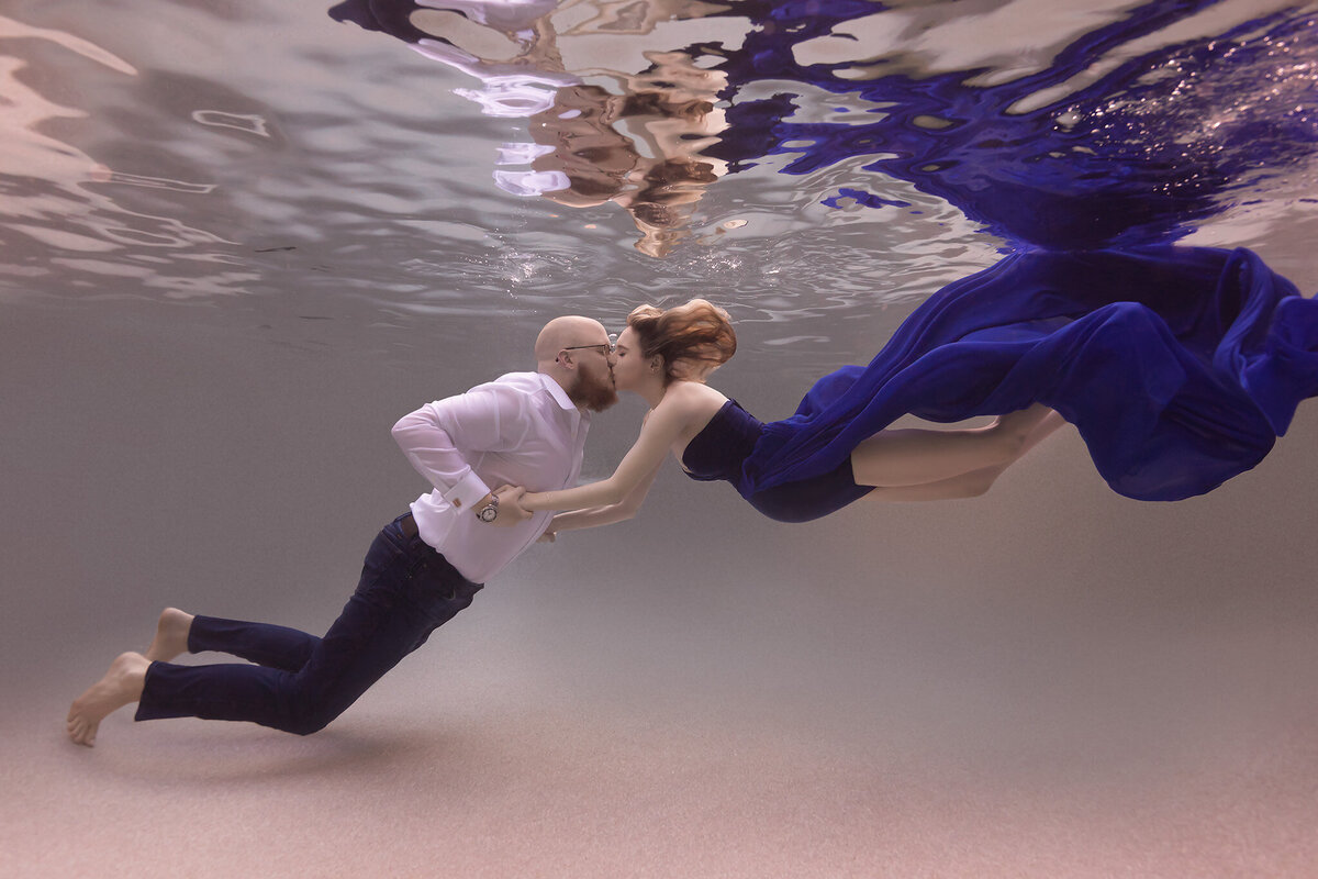  A captivating underwater maternity photo by a Dallas underwater photographer, featuring a couple sharing a tender kiss. The woman is dressed in a flowing blue gown, her hair drifting gracefully in the water, while her partner wears a crisp white shirt and dark pants. The tranquil water and the dreamy motion of the gown create a serene, romantic atmosphere.
