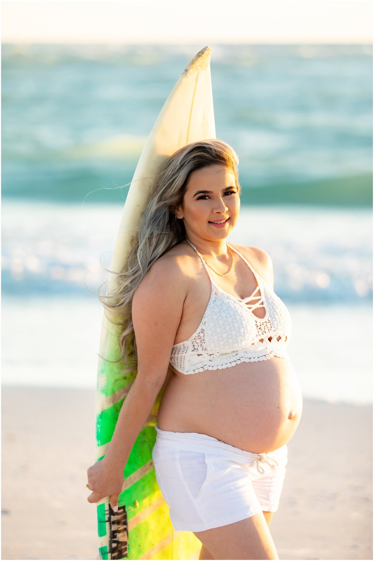 A maternity photoshoot with mom to be holding a surf board.