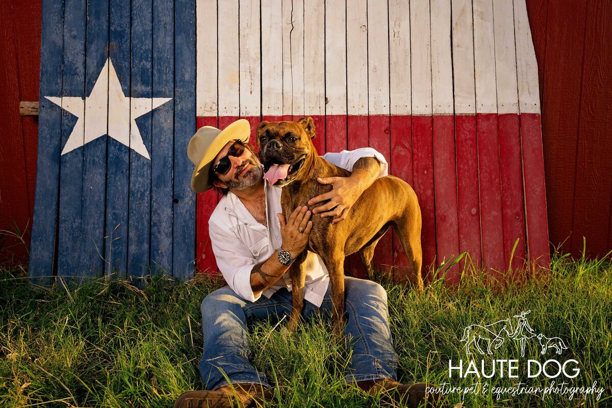 A man wearing sunglasses and a felt hat sits in front of a fence painted with the Texas flag and hugs his Boxer dog.