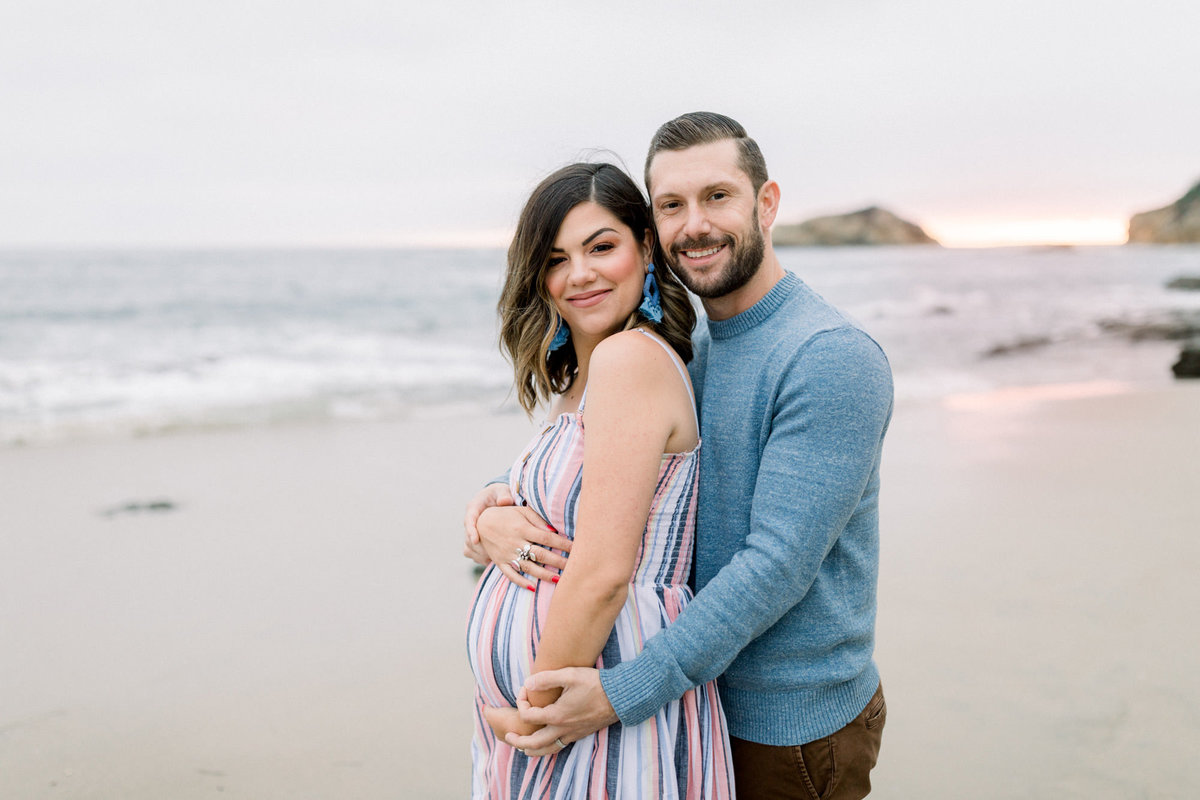 Soon to be dad embraces his wife from behind and holds her pregnant belly while at the beach