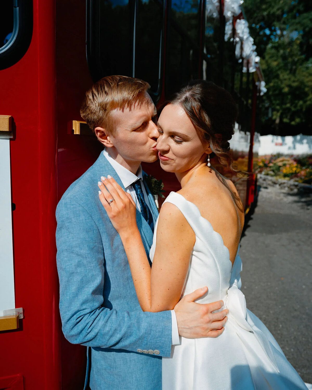 A groom in a blue suit kisses his bride on the cheek as they embrace beside a red vehicle, both smiling softly in bright outdoor sunlight—captured beautifully by a film photographer NJ couples trust.
