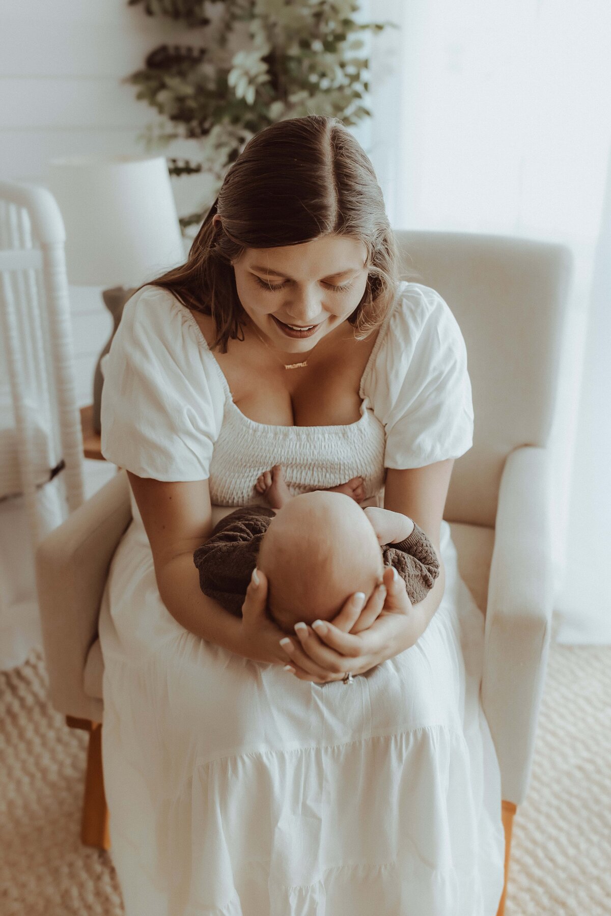 A woman in a white dress sits on a chair, smiling down at a baby cradled in her arms, in a softly lit room with plants in the background.