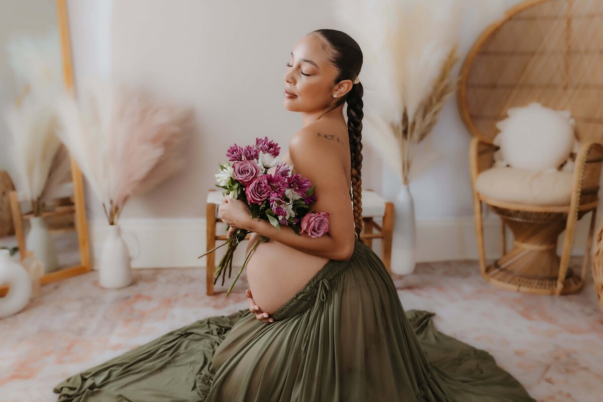 A pregnant woman with braided hair and a tattoo kneels in a room, holding a bouquet of pink and purple flowers. She is captured by a Macon maternity photographer in her flowing green dress, surrounded by pampas grass and a wicker chair.