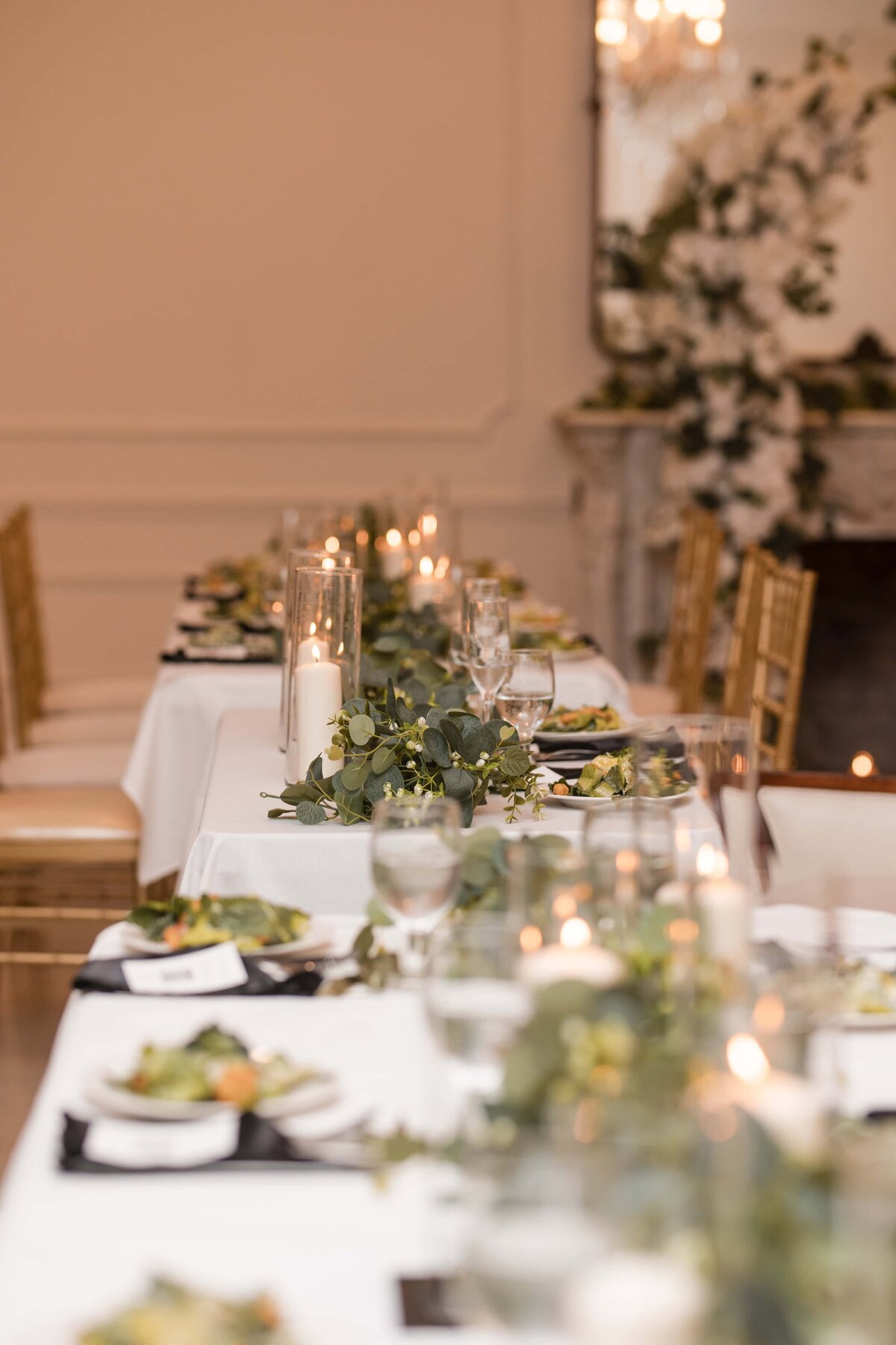 A beautifully styled wedding reception table at Azaleana Manor in Orange Park, Florida, featuring white linens, gold Chiavari chairs, greenery garlands, and glowing pillar candles in glass holders. Plates of salad are perfectly set for each guest, creating a warm and inviting atmosphere. The romantic and refined setup was captured by Heartful Films, a Jacksonville wedding photographer and videographer known for documenting the thoughtful details and elegant ambiance of wedding celebrations.