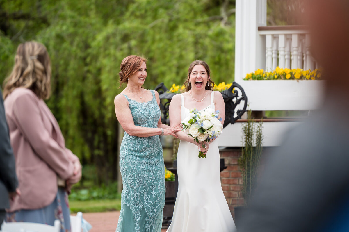 Two women standing outdoors, one in a white dress holding a bouquet, the other in a teal dress, both smiling and holding hands. People are gathered around them.