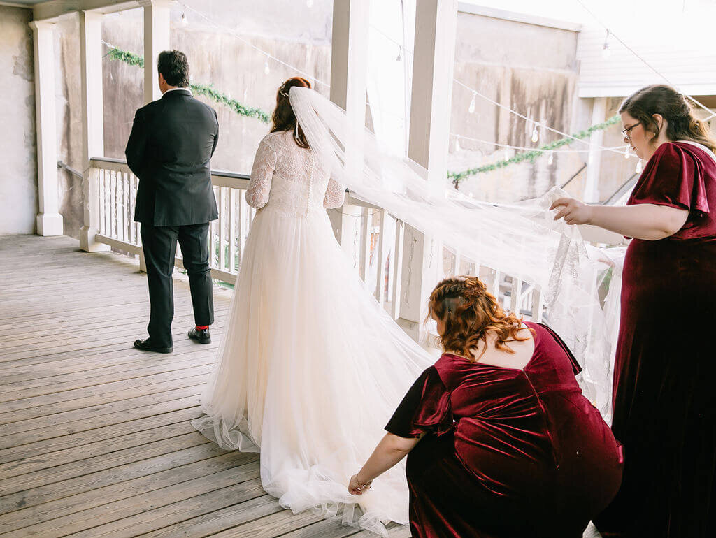 A bride stands behind a man in a black suite, facing away. Two bridesmaids in burgundy velvet dresses arrange the bride's dress and veil for a first look.