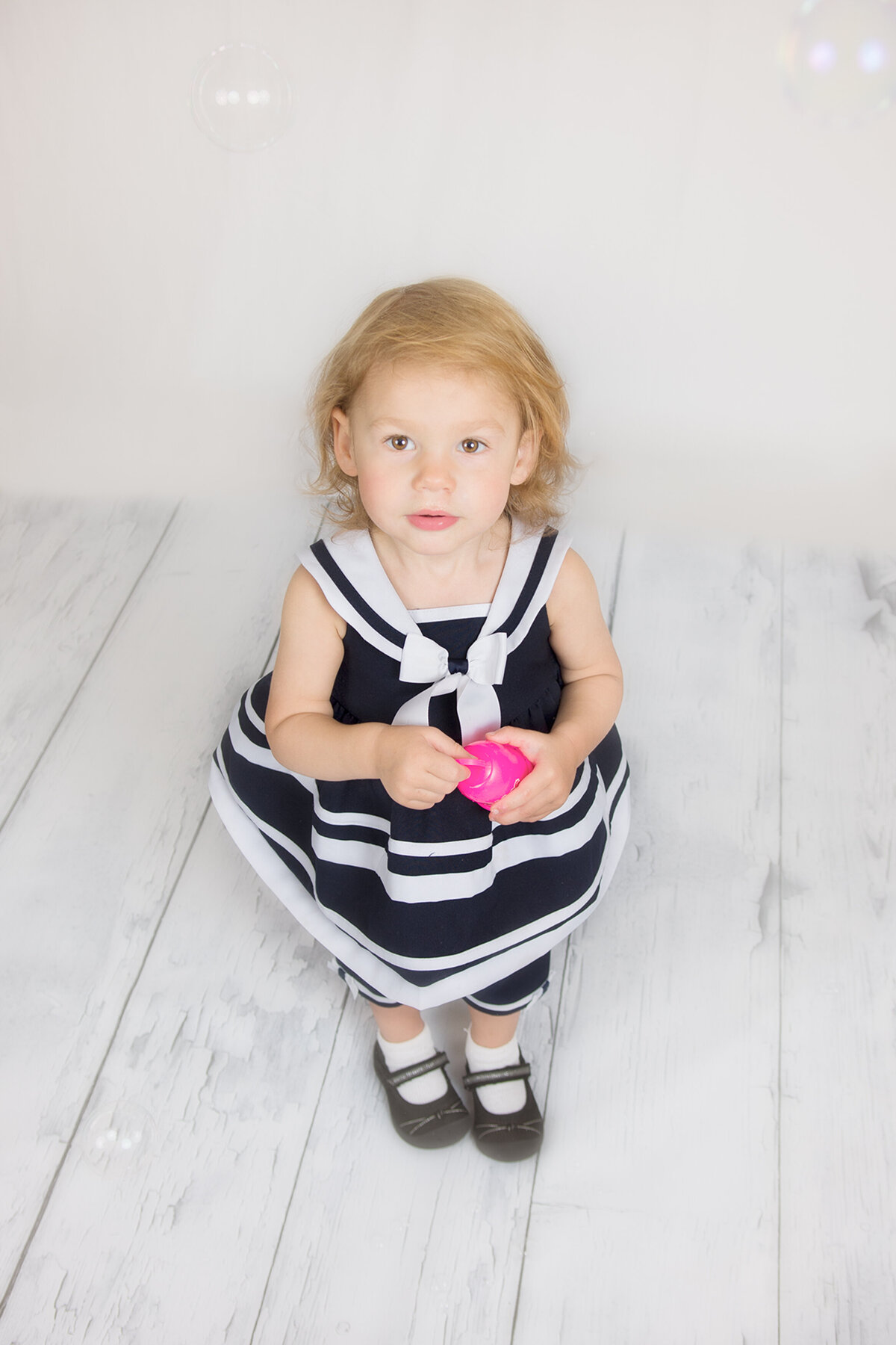 A toddler dressed in a Navy sailor outfit looking up at photographer Beth Benecki while she blows bubbles. It's studio-like setting with a white hardwood floor and white backdrop used for this family photo session.