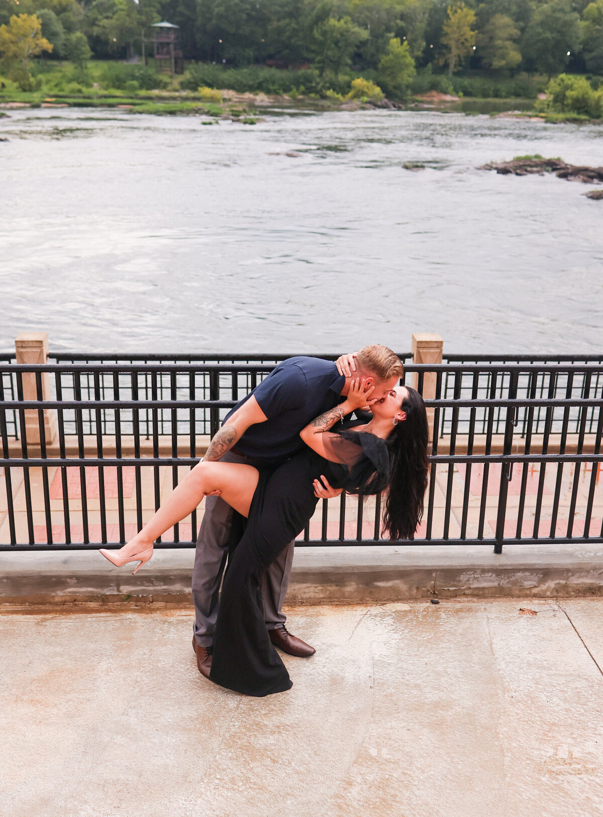 woman wearing black dress and man wearing gray pants and blue shirt dipping back kissing along brick walkpath with body of water behind them in columbus georgia
