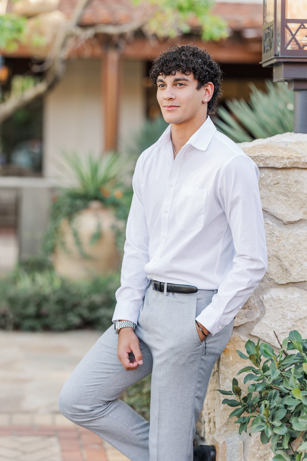 A confident senior guy poses at Mule Alley in Fort Worth, Texas, wearing a crisp white shirt and light gray slacks, leaning against a rustic stone wall with greenery in the background. Stylish and editorial portrait perfect for graduation photography.
