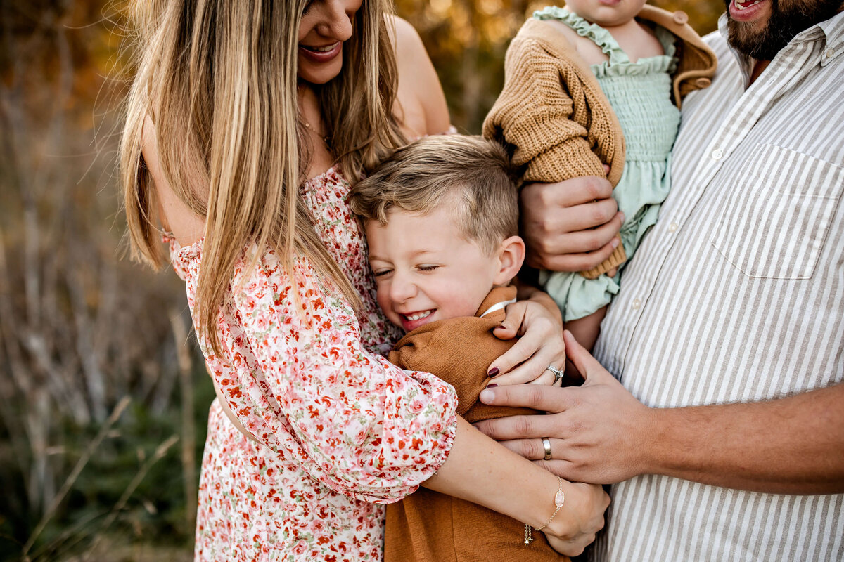 A woman hugs a smiling boy who seems delighted. A man and woman hold a baby in a green romper and brown sweater. The family is outdoors, and the scene conveys warmth and affection.