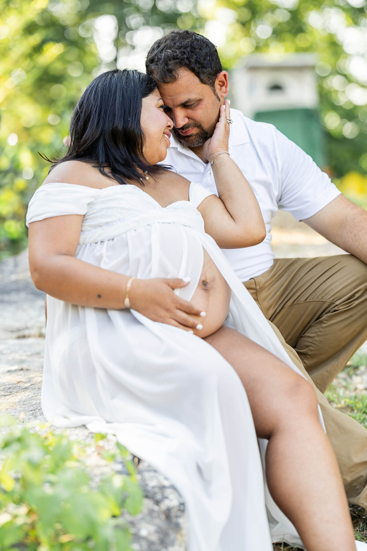 Expecting parents are sitting together cliff side with their heads together as the expecting woman holds her hand to the mans cheek 