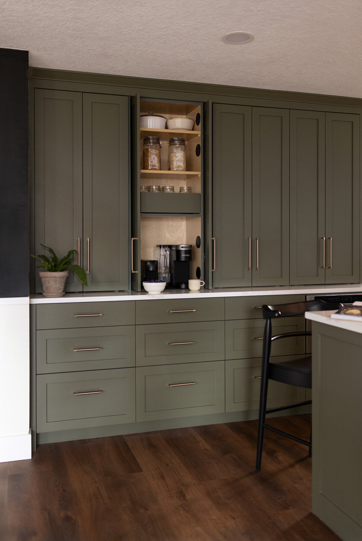 Kitchen with green cabinets, white countertops, and beige subway tile backsplash. Walnut floating shelves styled with books, plates, alcohol, and glasses. Black barstools at the island. Black pendant lights hang above the island.