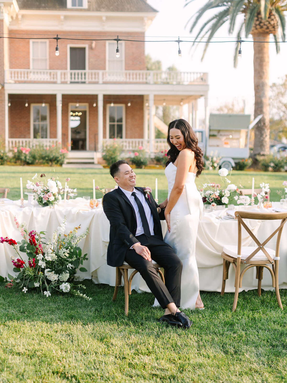 A smiling couple at an outdoor wedding reception. The man sits on a chair, and the woman stands beside him.
