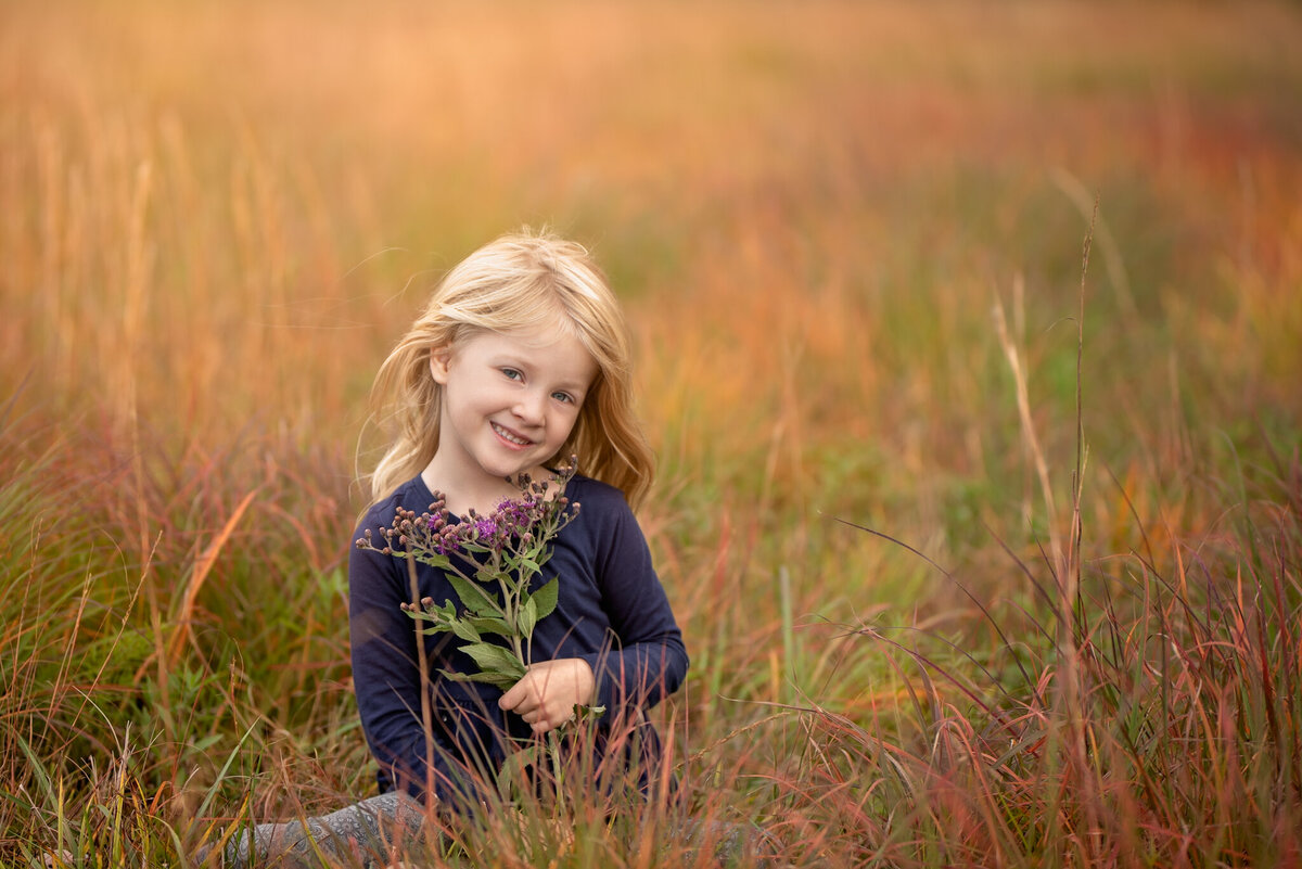 Candid portrait of a young girl standing in a sunlit meadow with vibrant grass. Captured in Gardner, Kansas, this natural scene is perfect for families looking for outdoor and relaxed child photography.