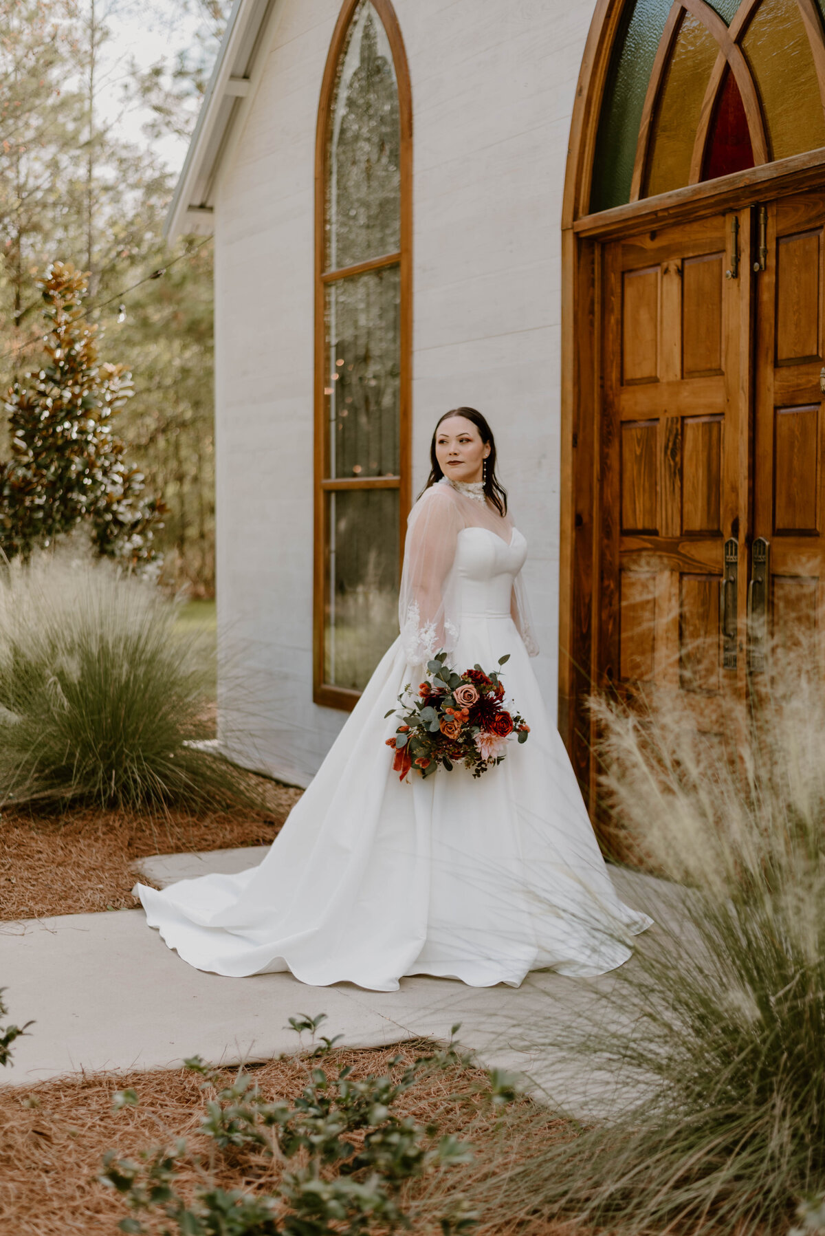 A portrait of a bride in her wedding dress holding her bouquet standing in front of a stained glassed door at the cypress grove open air chapel.