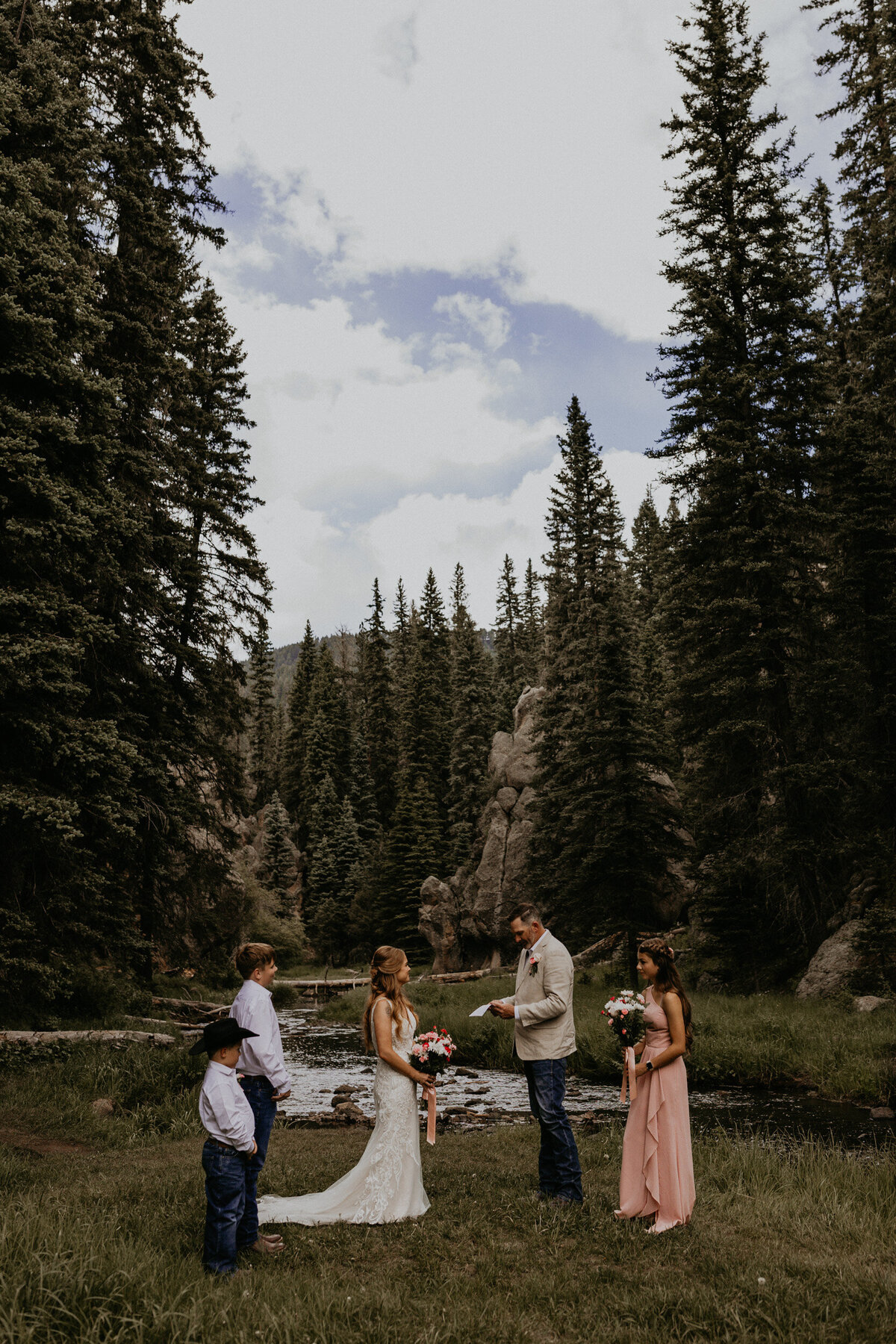 a bride and groom standing together during their intimate wedding ceremony in front of a creek with their kids