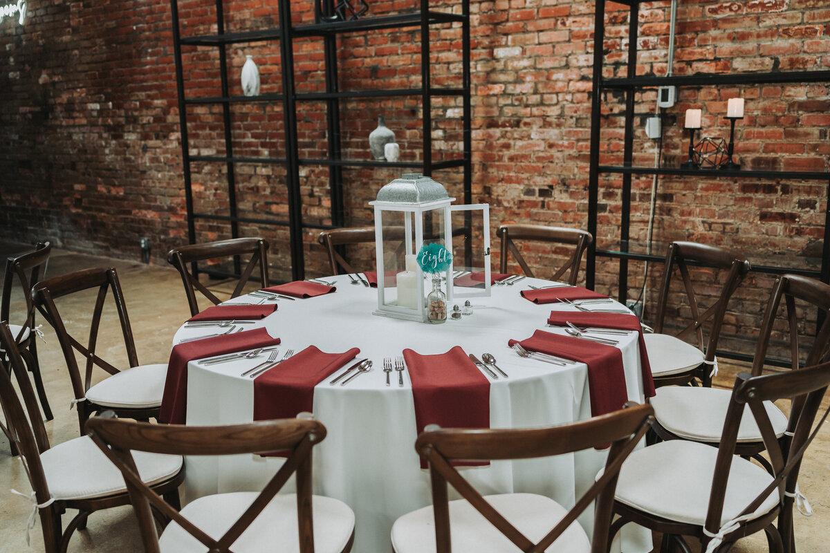 An elegant wedding reception table set with white linen, burgundy napkins, and a white lantern centerpiece, surrounded by dark wooden chairs, against an industrial brick wall at The Neon House in Norfolk, Virginia, captured by Quinn Photography LLC.