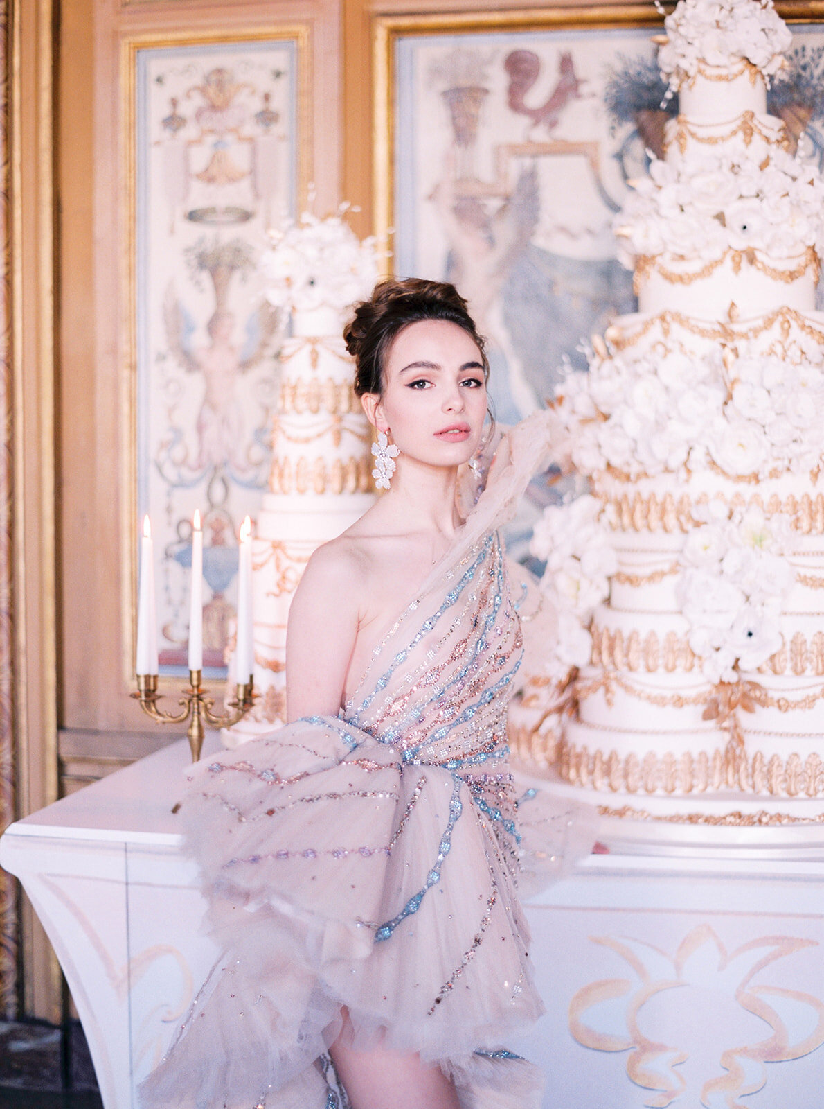 The exquisite photo was captured at the stunning Chateau Vaux le Vicomte in Paris, France, known for its enchanting beauty and grandeur, making it one of the top destination wedding venues in the country. Photographer Audrey FORA expertly framed the scene, showcasing a woman in a breathtaking, one-shoulder gown embellished with sequins and ruffles. She stands elegantly before an opulent multi-tiered cake adorned with delicate white flowers, set against a backdrop of ornate, gold-accented walls and a gleaming candelabra, adding a touch of romance to the luxurious ambiance. Additionally, Videographers East West Films were present to capture the magical moments on film, ensuring every detail was preserved for eternity.
