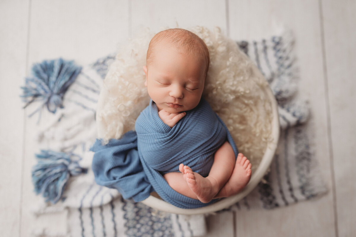 Newborn boy in blue swaddle with feet hanging out lying in white bowl placed on top of blue striped rug